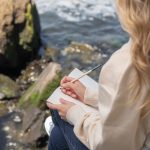 woman sitting on a rock by the water writing in her journal